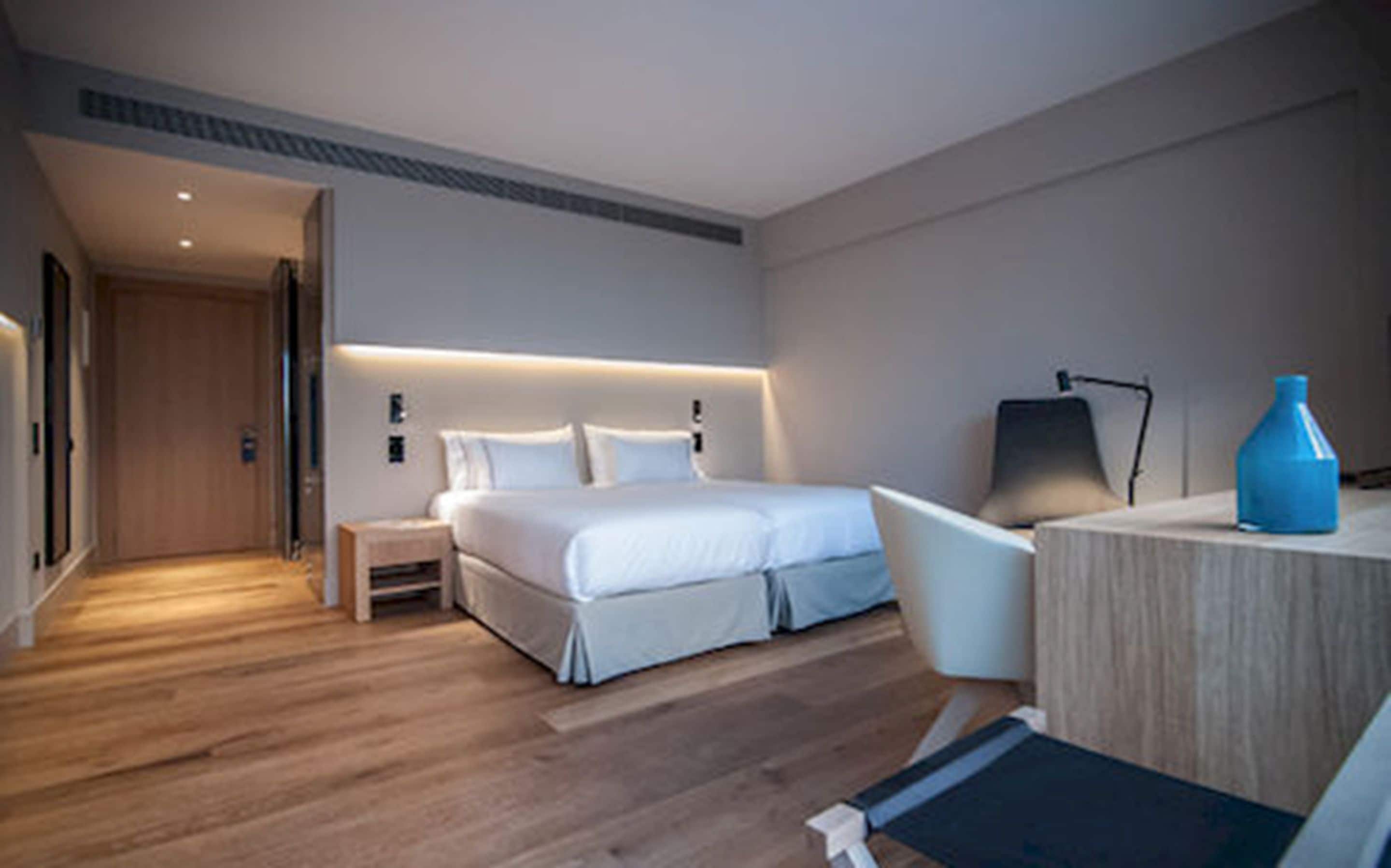 A modern hotel room with a wooden floor, a bed with white bedding, a desk with a chair, and a blue vase on the desk.