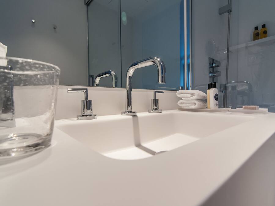 Un baño moderno con un lavamanos blanco, tres grifos cromados y un cuenco de vidrio sobre la encimera.