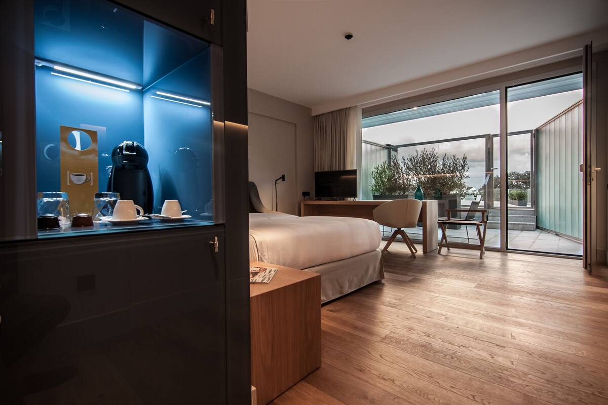 A modern hotel room with a large bed, a desk, a chair, a TV, and a balcony with a view.