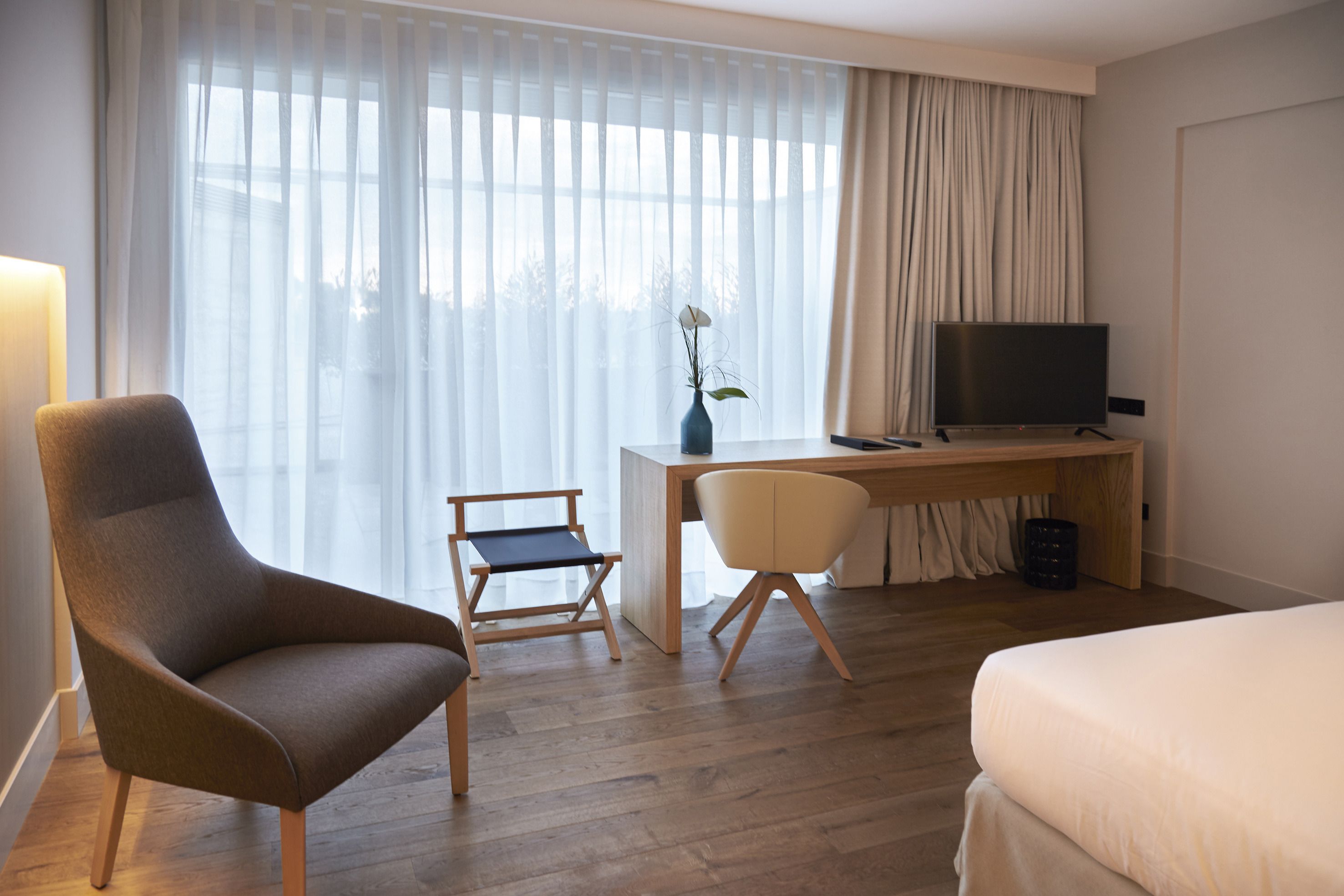 A hotel room with a bed, a desk with a television, a chair, and a vase with a flower.