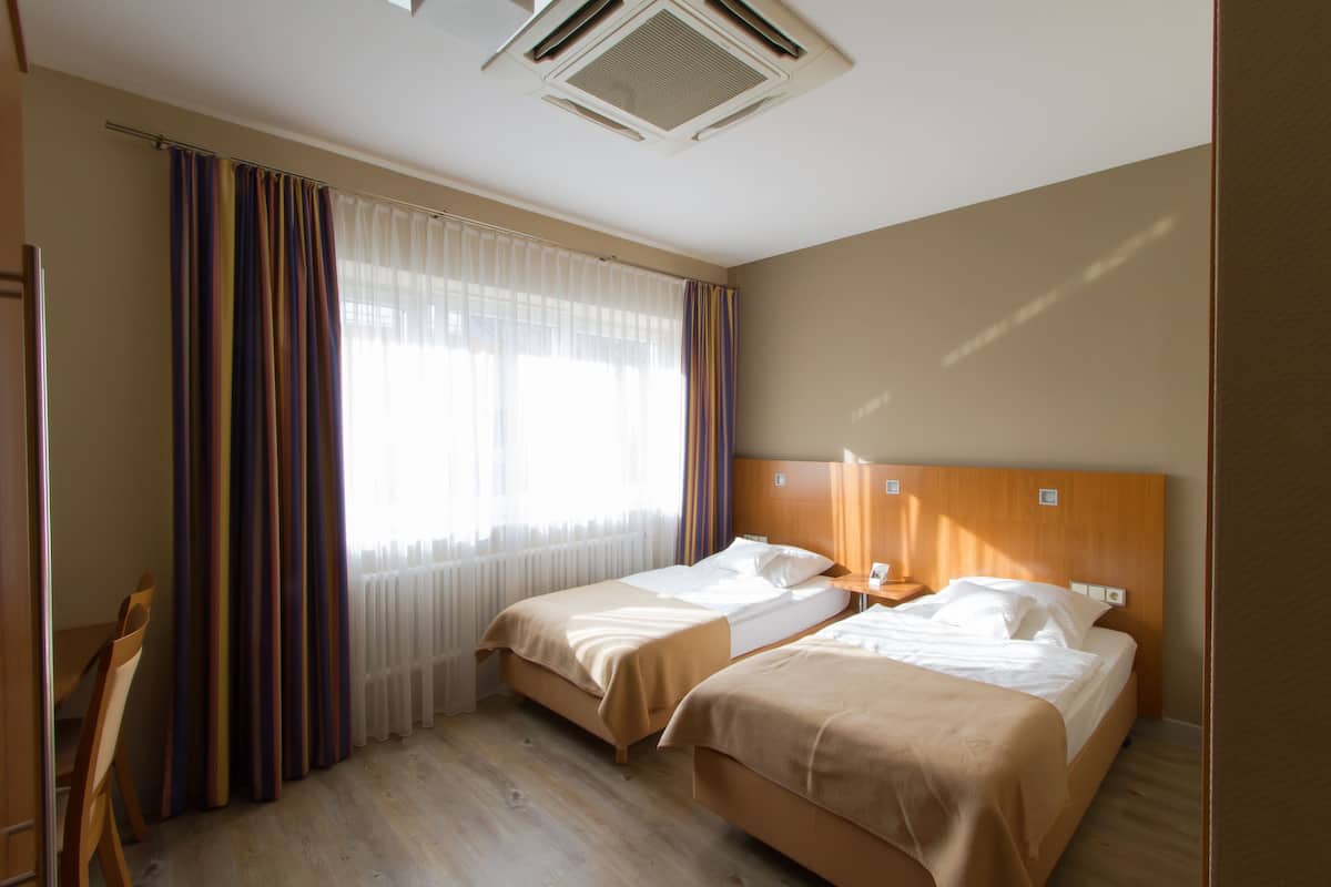 Hotelkamer met twee bedden, een houten hoofdbord, een raam met gordijnen, een radiator en een houten stoel.