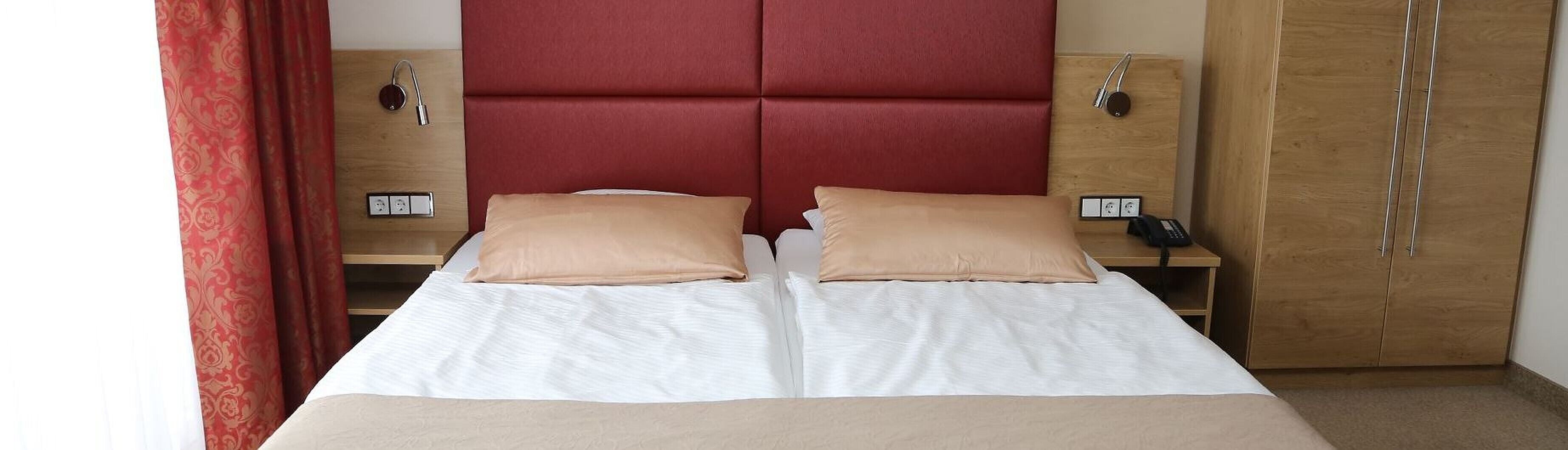 Un lit double avec deux oreillers beige, une tête de lit rouge, des tables de chevet avec des lampes et une armoire en bois.