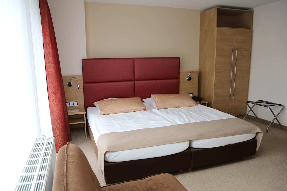 Een net opgemaakt bed met een rood hoofdeinde, een houten garderobe en een klein nachtkastje met een lamp.