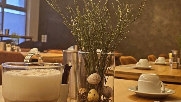 A dining table with a glass of frothy beverage, a clear glass vase with dried branches and eggs, and a cup and saucer in the background.