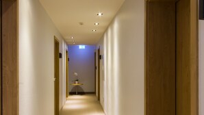 A narrow, well-lit hallway with wooden doors on both sides, leading to a room with a small table and a potted plant.