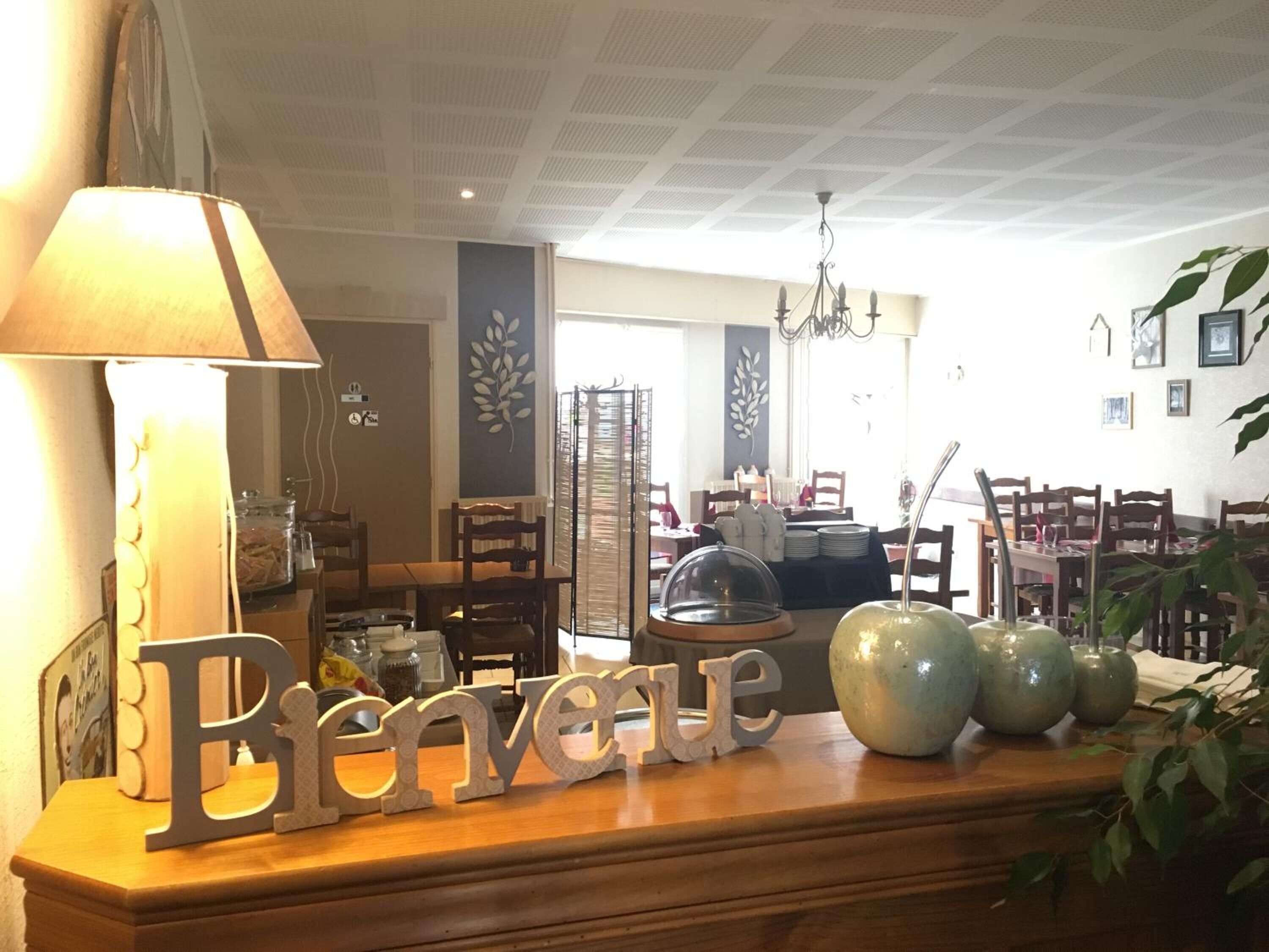 Un hall de hotel con un cartel de 'Bienvenue', una lámpara, una araña y un área de comedor al fondo.