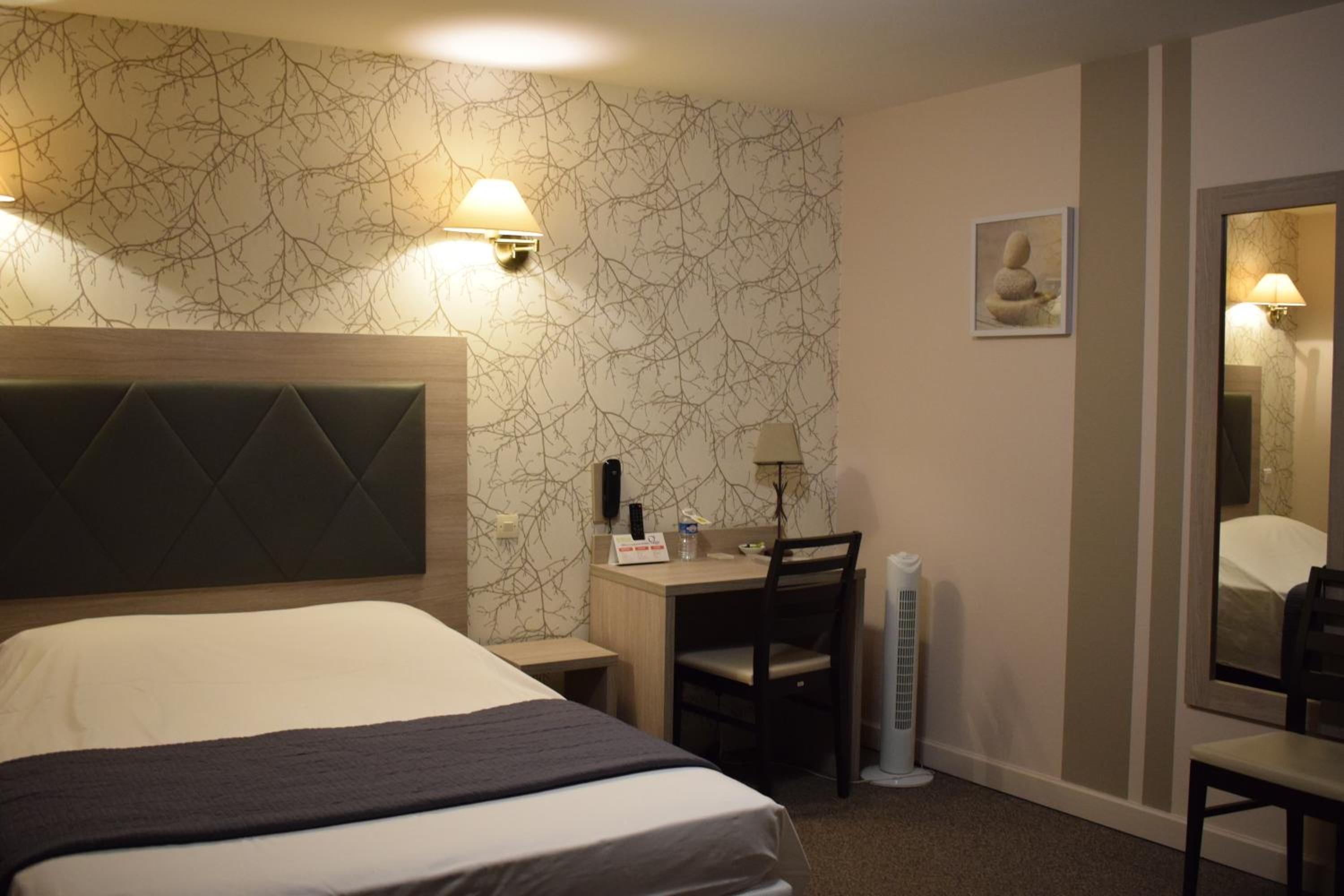 A hotel room with a large bed, a desk with a chair, a standing heater, and a wall-mounted lamp.