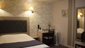 A hotel room with a large bed, a desk with a chair, a standing heater, and a wall-mounted lamp.