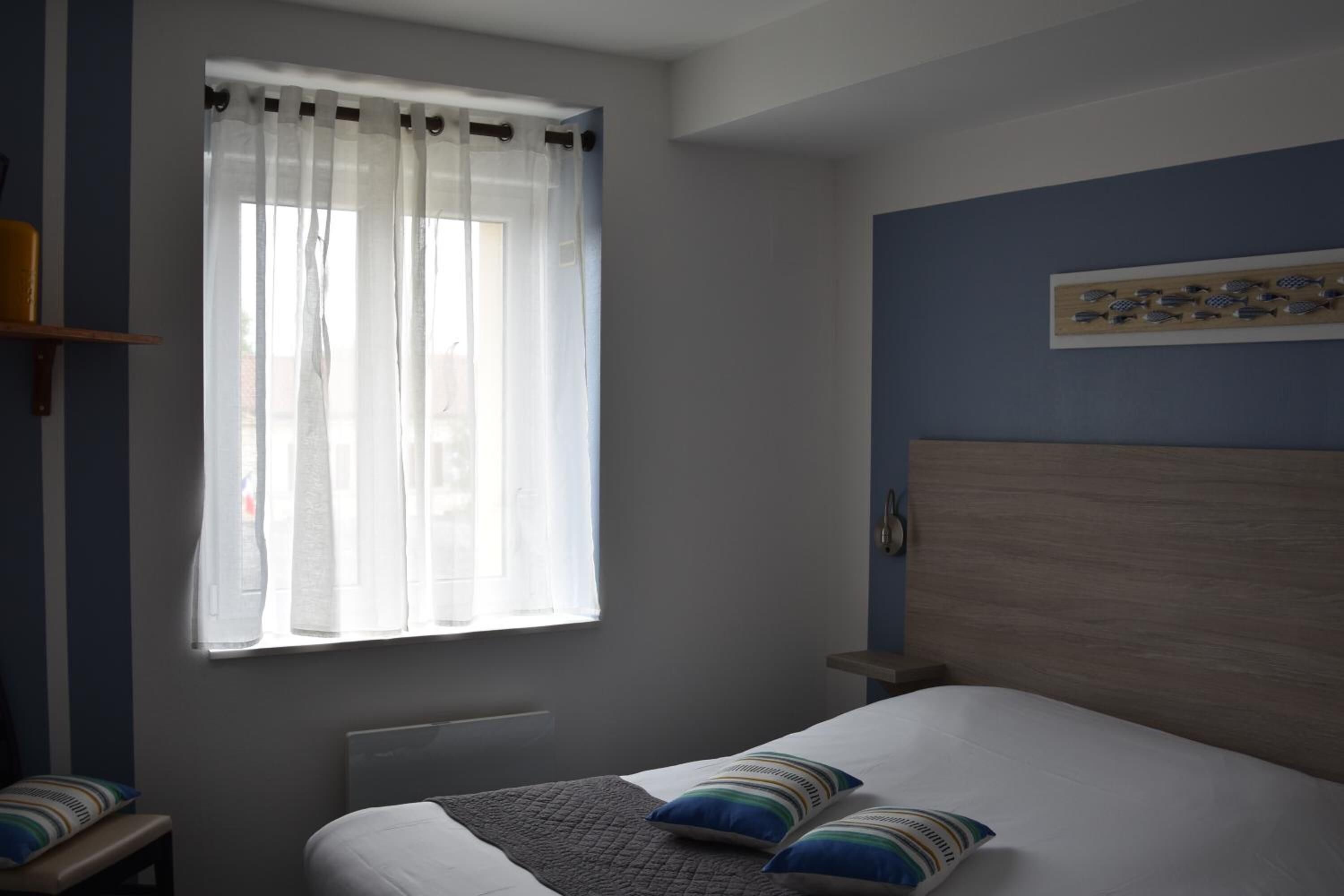 A bedroom with a bed, a window with sheer curtains, a bedside table, and a wall-mounted shelf.