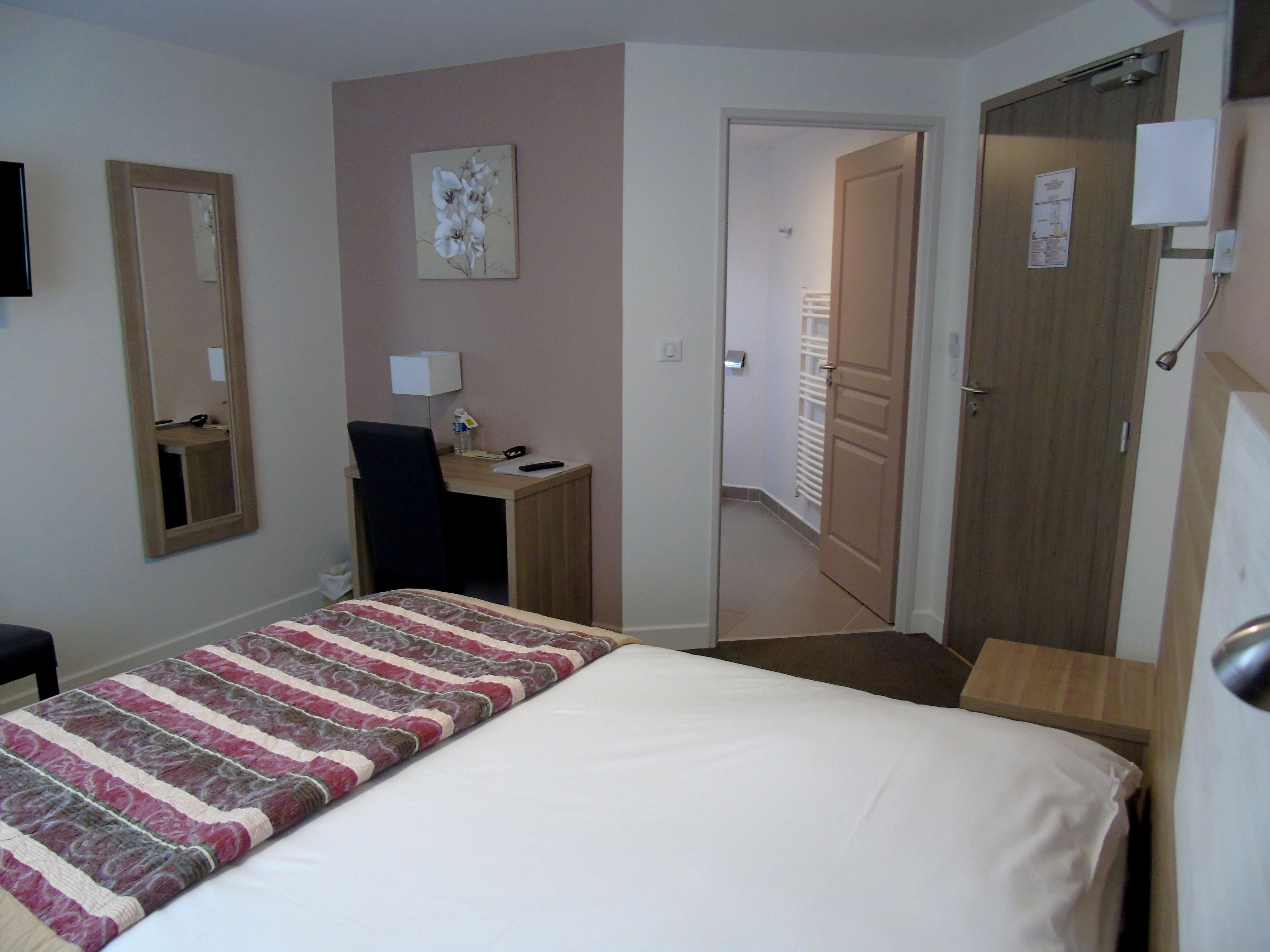 A hotel room with a bed, a desk, a chair, a mirror, and a door leading to another room.