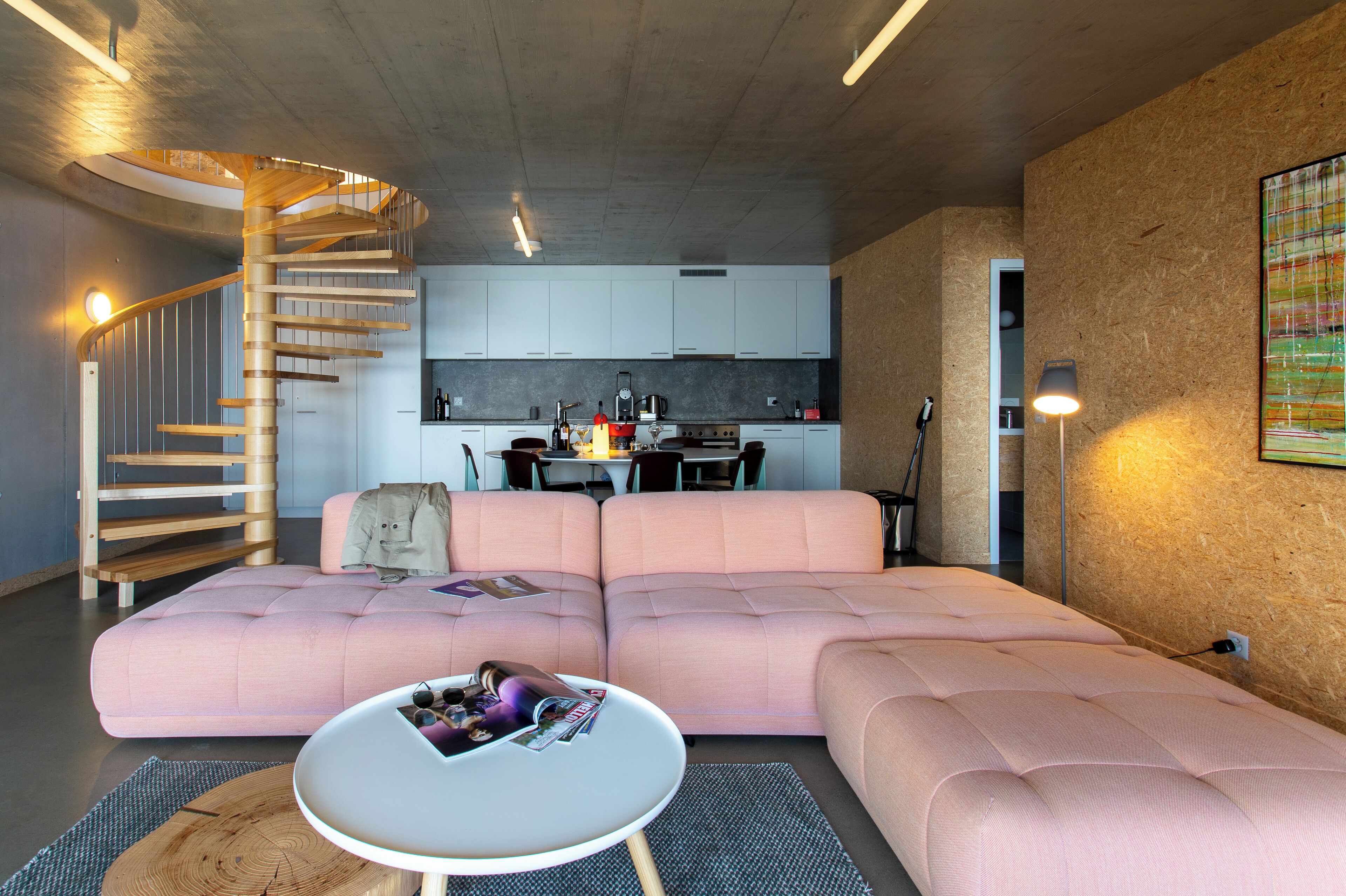 Un salon moderne avec un canapé d’angle rose, une table basse ronde et un escalier en colimaçon.