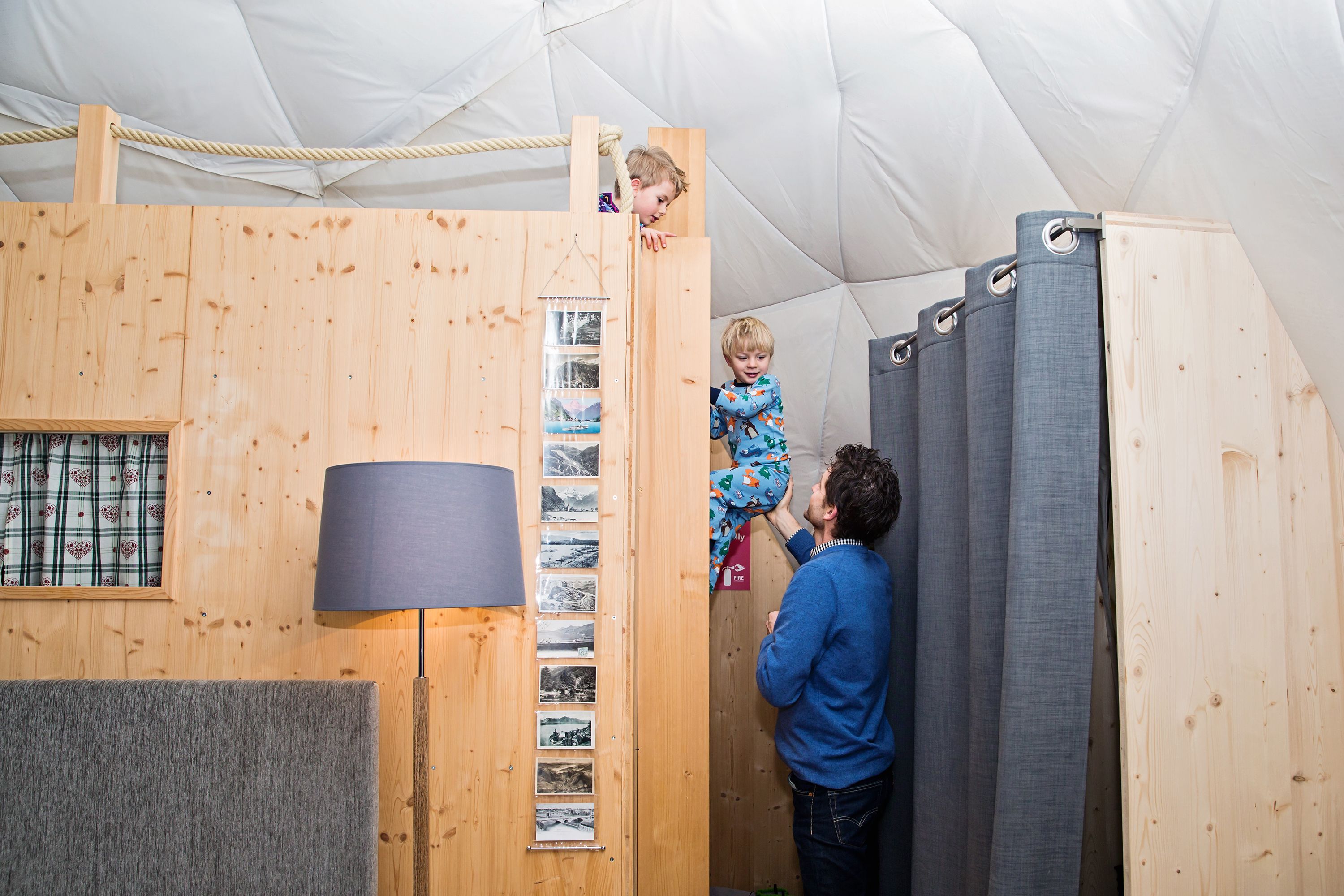 A wooden play structure with a ladder, a wall-mounted lamp, and a window with patterned curtains.