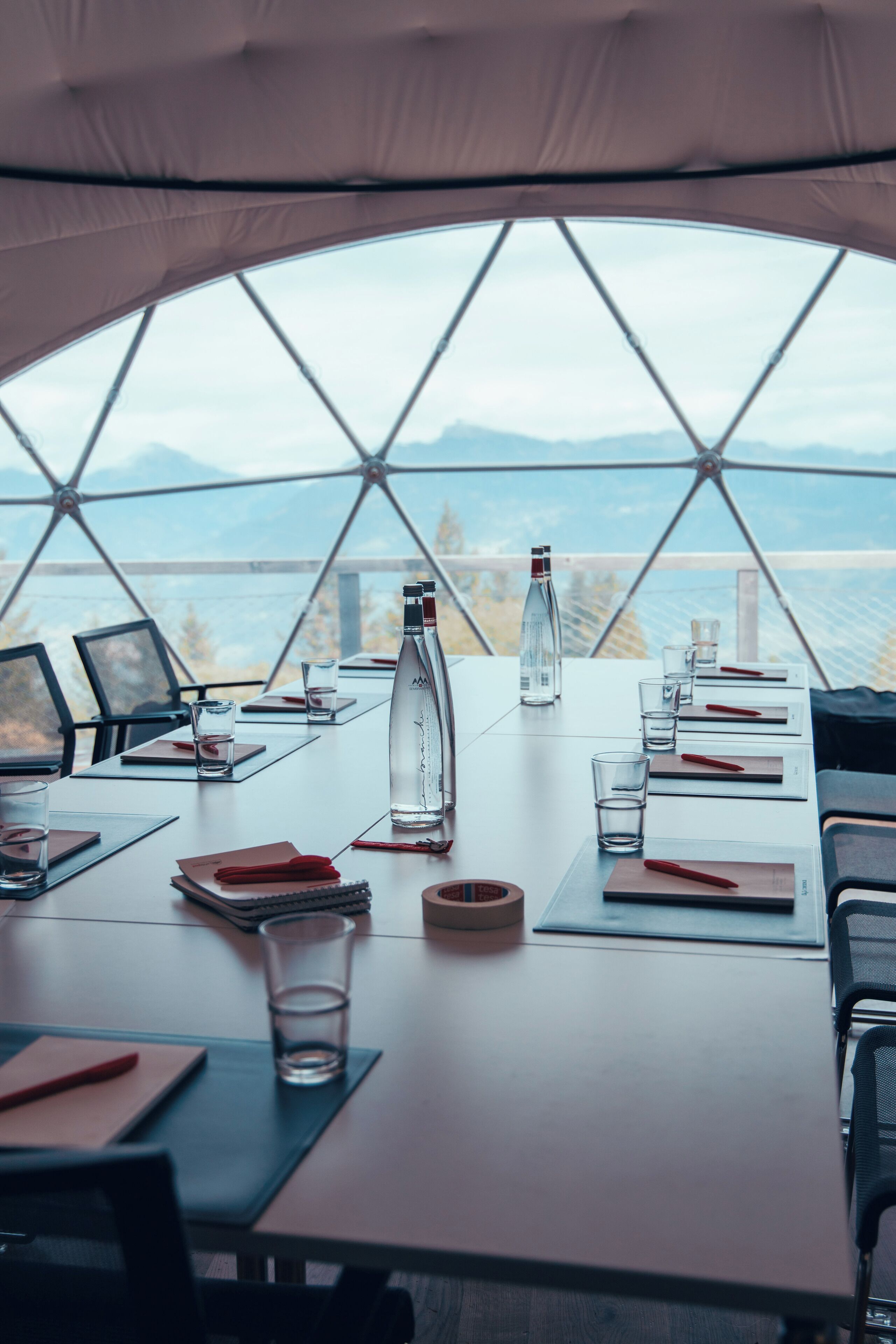 A dining table set up inside a geodesic dome with a panoramic view of mountains and a lake.