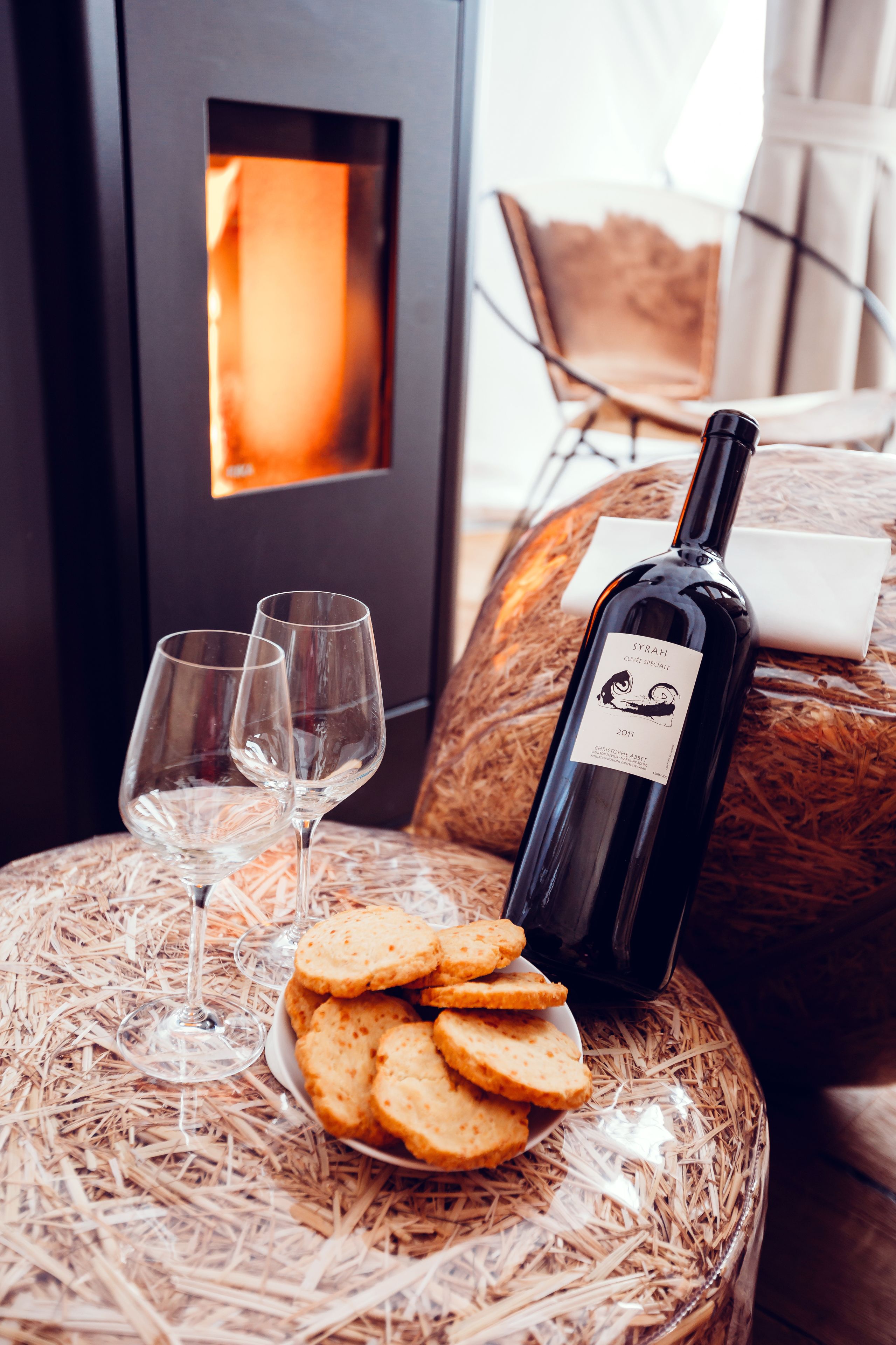 A cozy room with a fireplace, two wine glasses, a bottle of wine, and a plate of cookies on a woven stool.