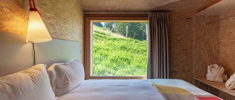 A hotel room with a large bed, a window with a view of a green hillside, a bedside lamp, and a shelf with towels.