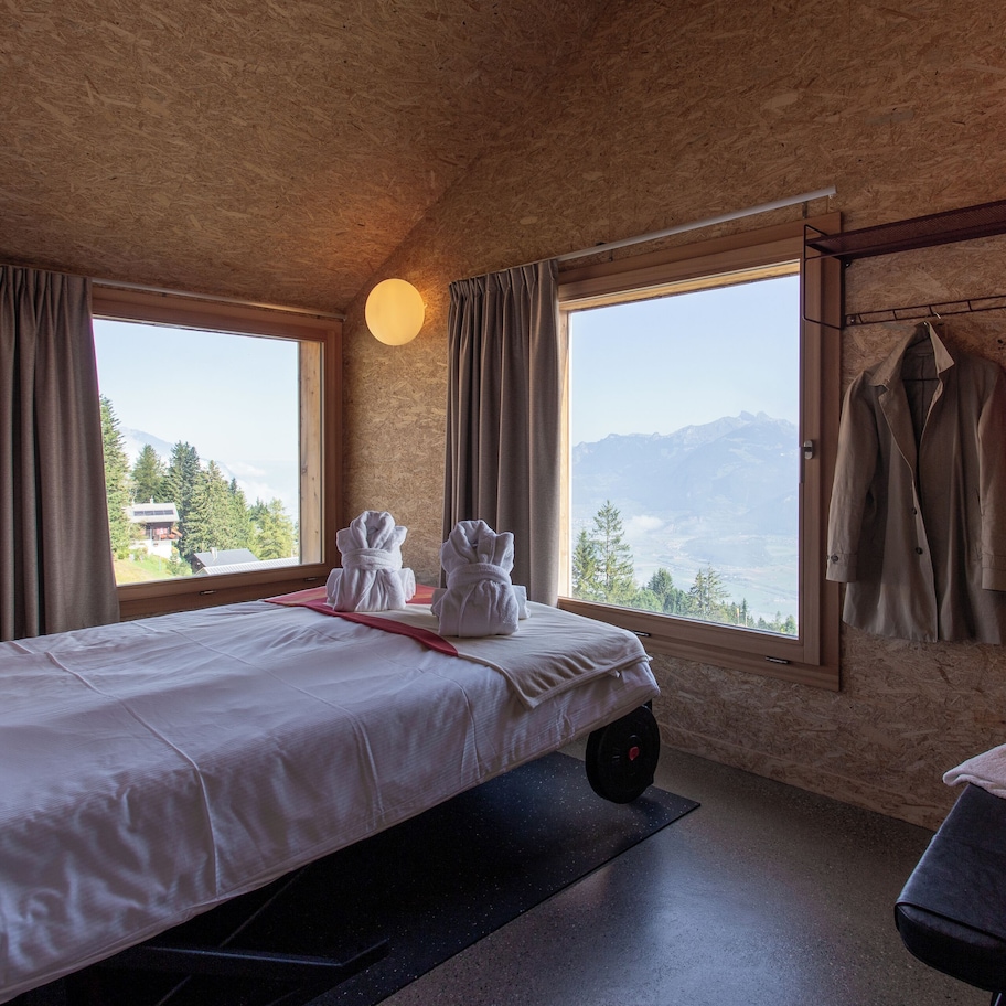 A room with a bed, a bench, and a view of mountains.