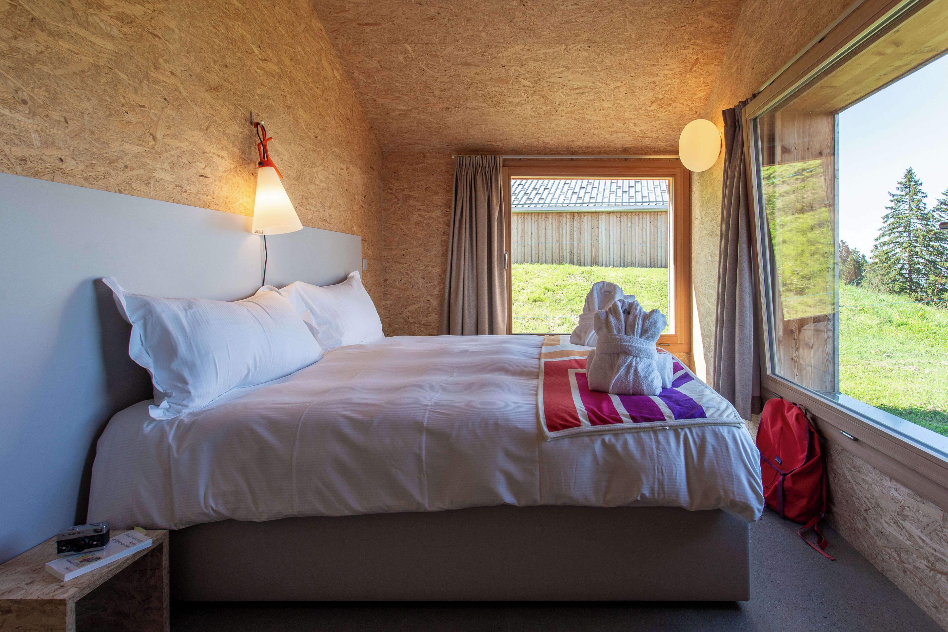 A bedroom with a large bed, a nightstand, a lamp, a window with curtains, and a view of a grassy area.