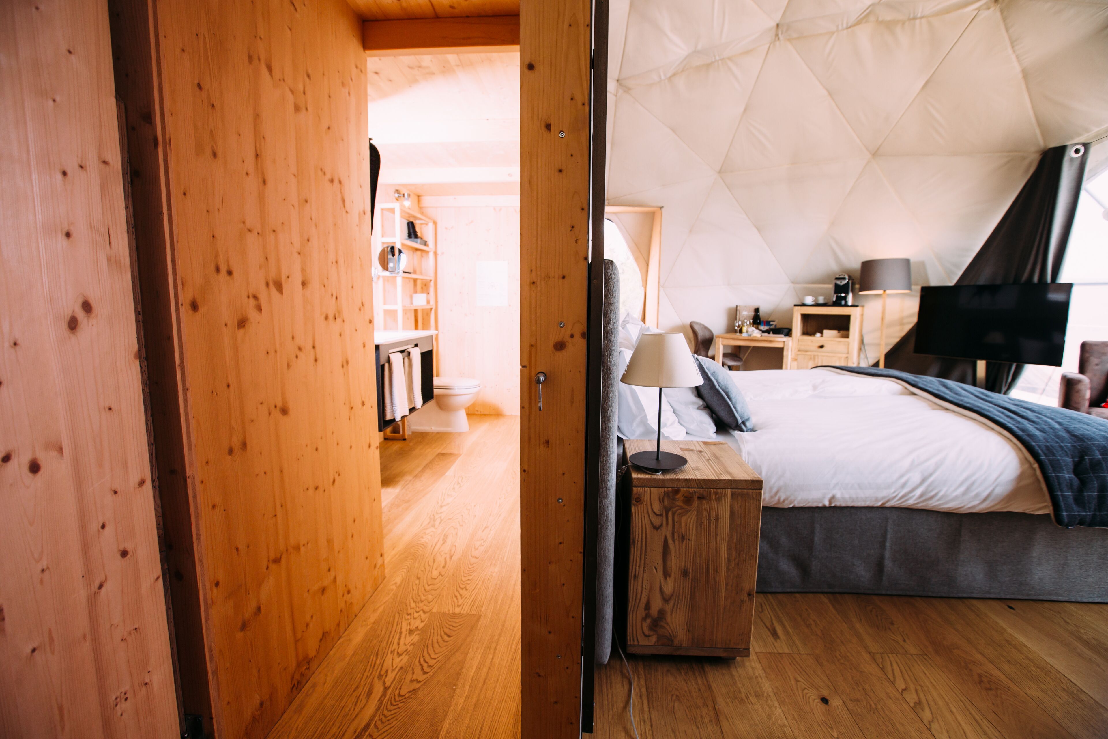 Ein gemütliches Schlafzimmer mit Holzboden, einem Bett mit grauer Decke, einem Nachttisch mit Lampe und einem Fernseher auf einem Schrank.