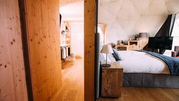 Ein gemütliches Schlafzimmer mit Holzboden, einem Bett mit grauer Decke, einem Nachttisch mit Lampe und einem Fernseher auf einem Schrank.