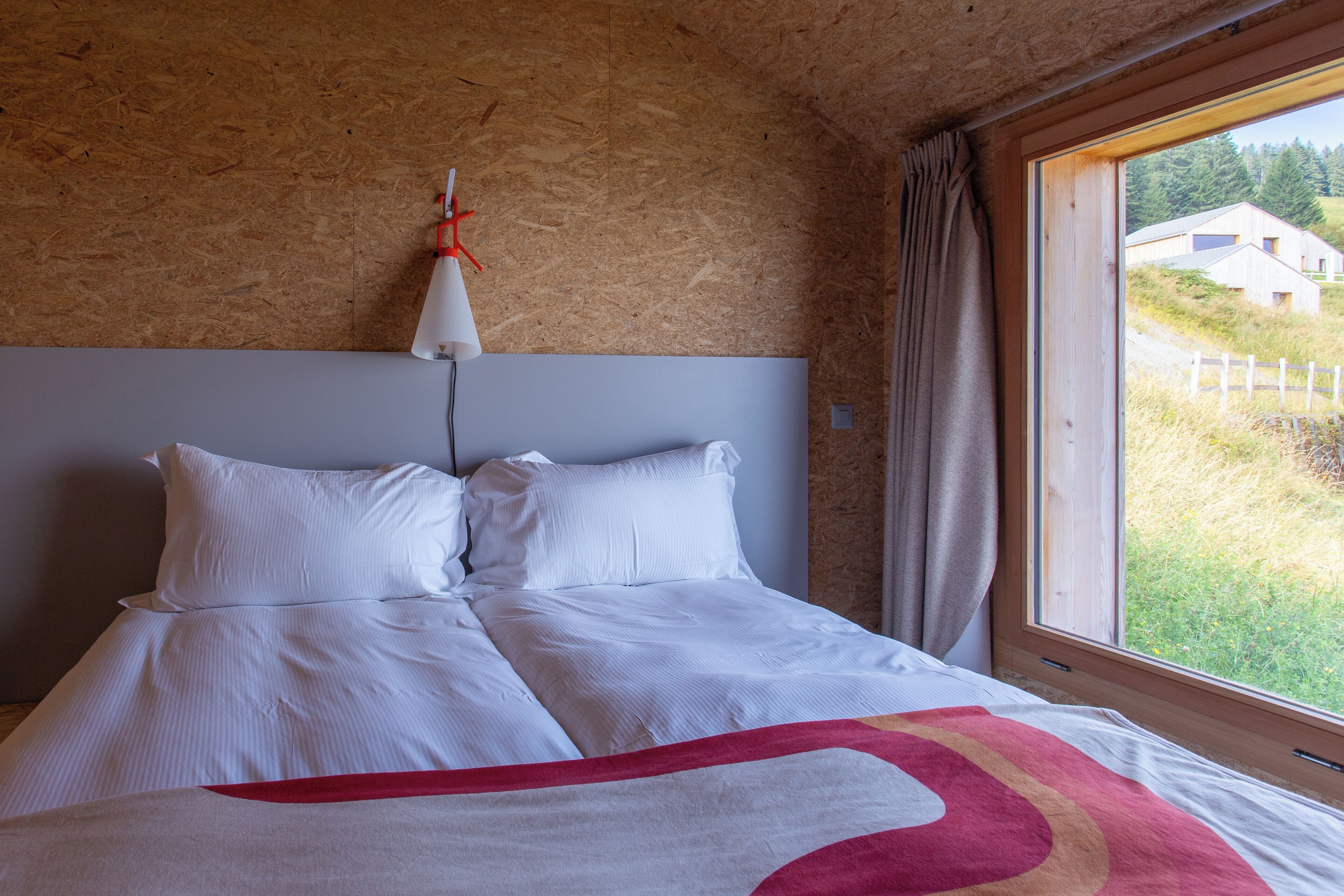 Een slaapkamer met een groot bed, een houten hoofdbord, een raam met uitzicht op een huis en bomen, en een gordijn.