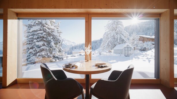 Una habitación con un ventanal grande que da a un paisaje nevado, dos sillas y una mesa con un florero y vasos.