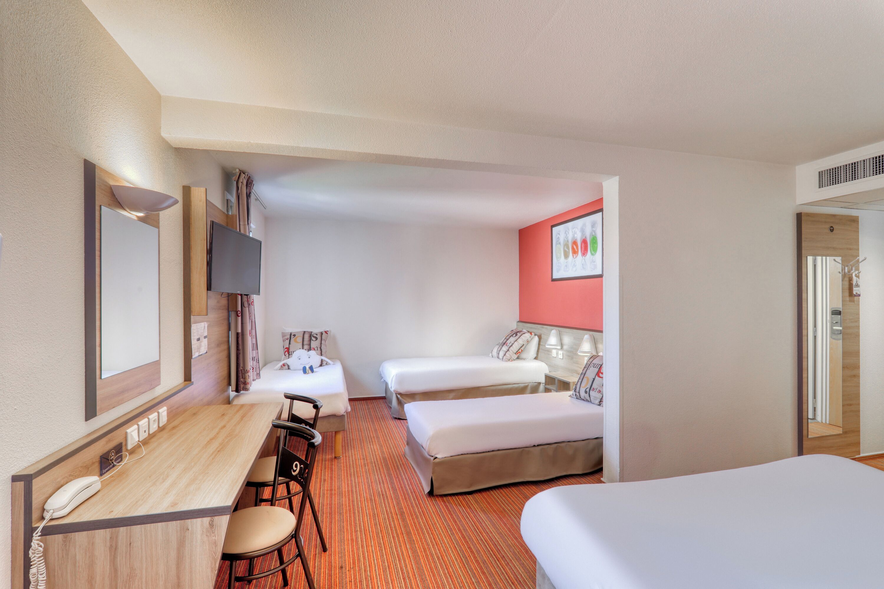 A hotel room with a desk, two beds, a TV, and a red accent wall.