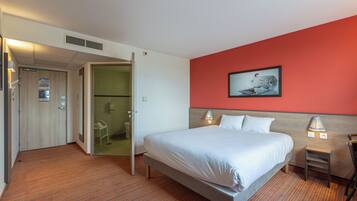 Una habitación de hotel con cama, una silla, un escritorio y un baño visible a través de una puerta abierta.