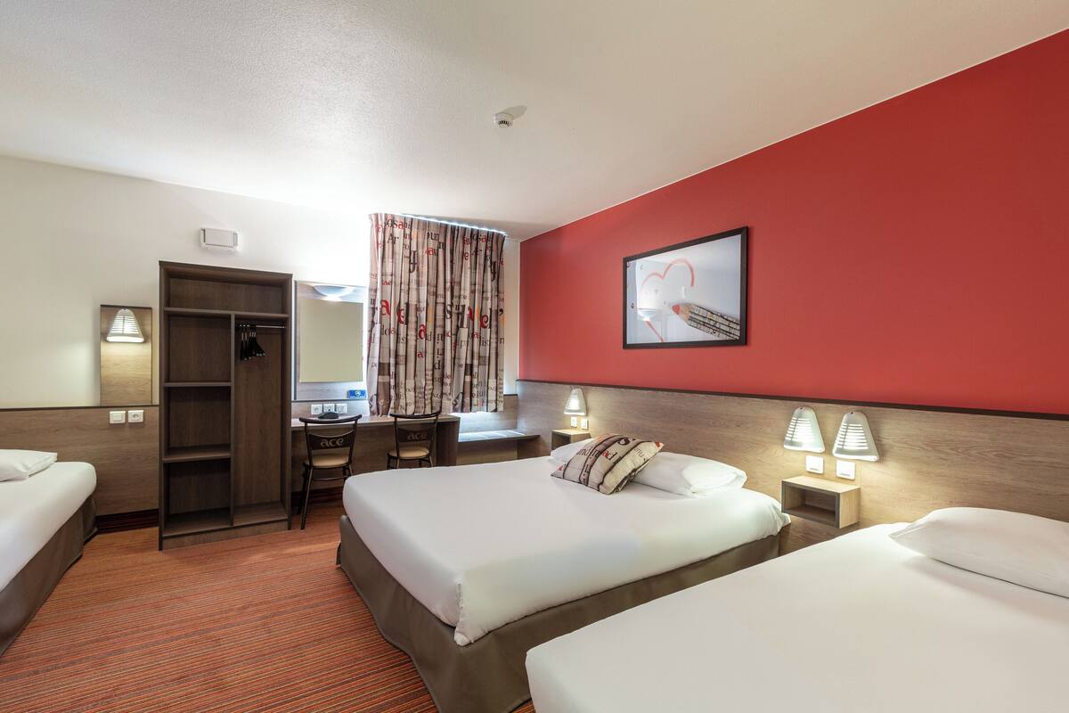 Habitación de hotel con dos camas, un escritorio, una silla, un armario y una pared roja decorativa.