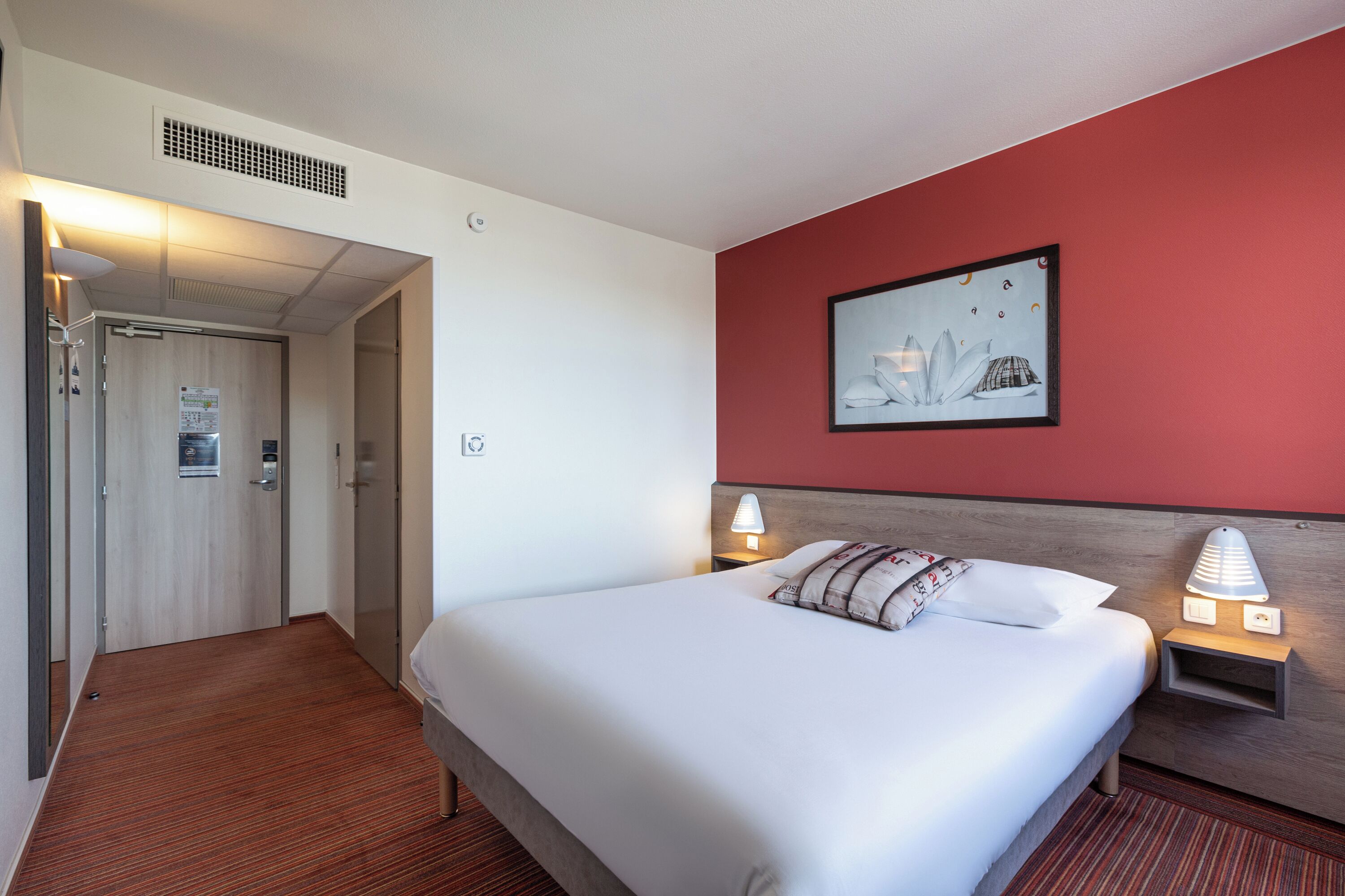 Une chambre d’hôtel avec un lit, des lampes de chevet, un mur d’accent rouge, un tableau encadré et une porte donnant sur une autre pièce.