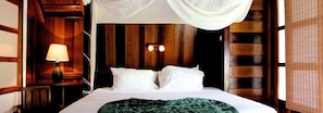 A bed with white linens and a green patterned blanket, a bedside lamp, and wooden paneling on the walls. - Acajou Hotel (Grande Riviere)