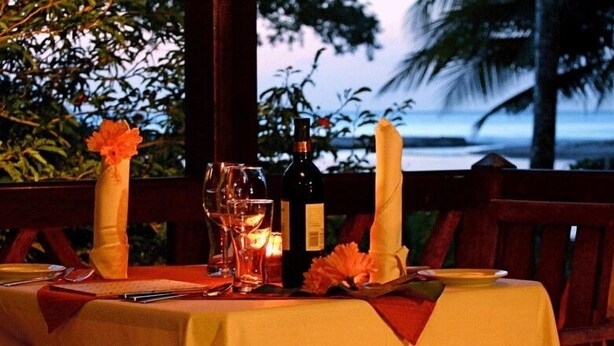 A table set for two with a bottle of wine, glasses, and a candle, overlooking a beach at dusk. - Acajou Hotel (Grande Riviere)