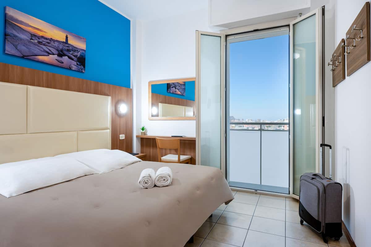 A hotel room with a bed, a desk, a chair, a window, and a balcony.