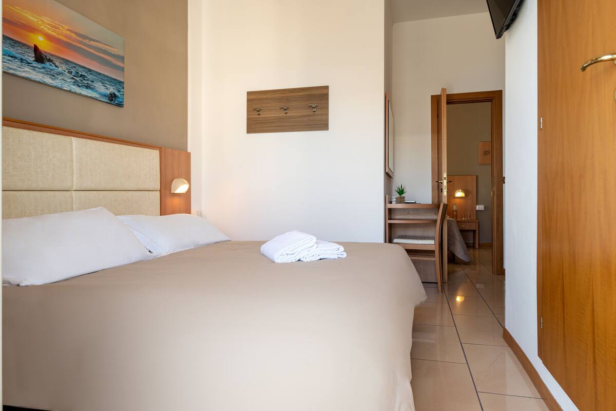 A hotel room with a large bed, a wooden headboard, a painting of a sunset over the ocean, a wooden panel on the wall, and a wooden door.