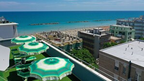 A rooftop terrace with green and white striped umbrellas, a view of the sea, and buildings in the background. - Amadei Hotel Figaro & Apartments (Pesaro)