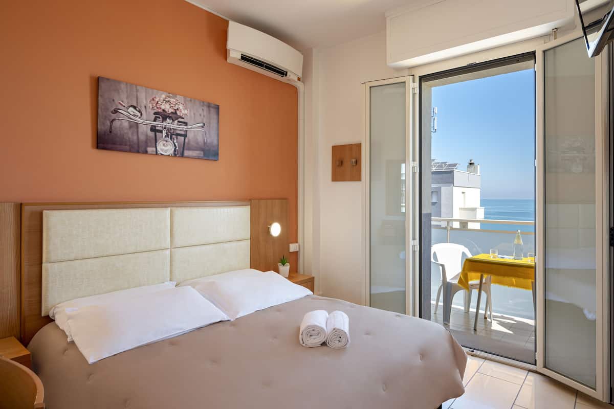 A hotel room with a bed, a painting on the wall, a small table with a yellow tablecloth, and a view of the sea through a sliding glass door.
