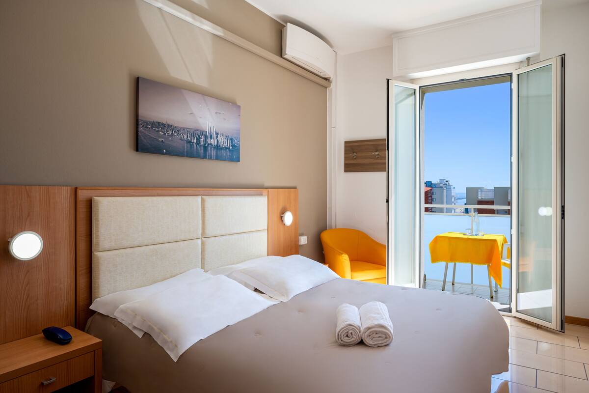 A hotel room with a bed, a bedside table, a chair, and a balcony with a table.