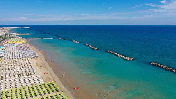 An aerial view of a beach with rows of umbrellas and sun loungers, a clear blue sea, and a rocky breakwater. - Amadei Hotel Figaro & Apartments (Pesaro)