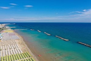 Vista aérea de una playa con hileras de sombrillas y tumbonas, un mar azul transparente y un rompeolas rocoso.