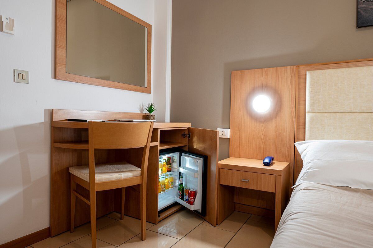 Une chambre d’hôtel compacte comprenant un lit, un bureau avec une chaise, un petit réfrigérateur, une table de chevet avec une lampe et un miroir au mur.