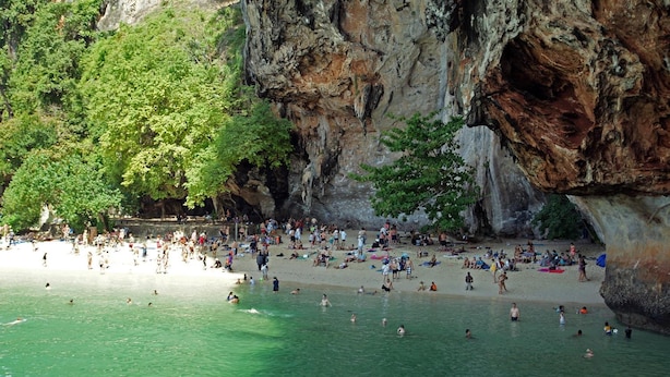 A crowded beach with people swimming and sunbathing, surrounded by rocky cliffs and lush greenery.