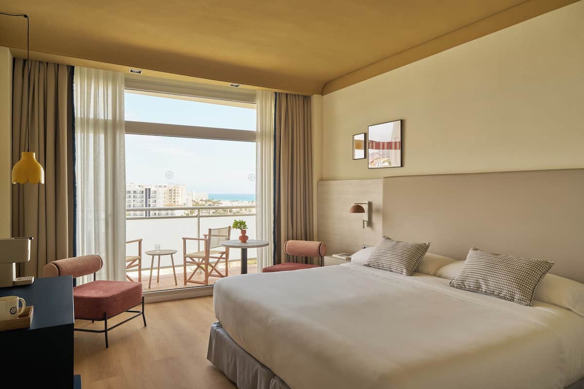 A hotel room with a large bed, a desk, a chair, a small table, and a view of the city through large windows.