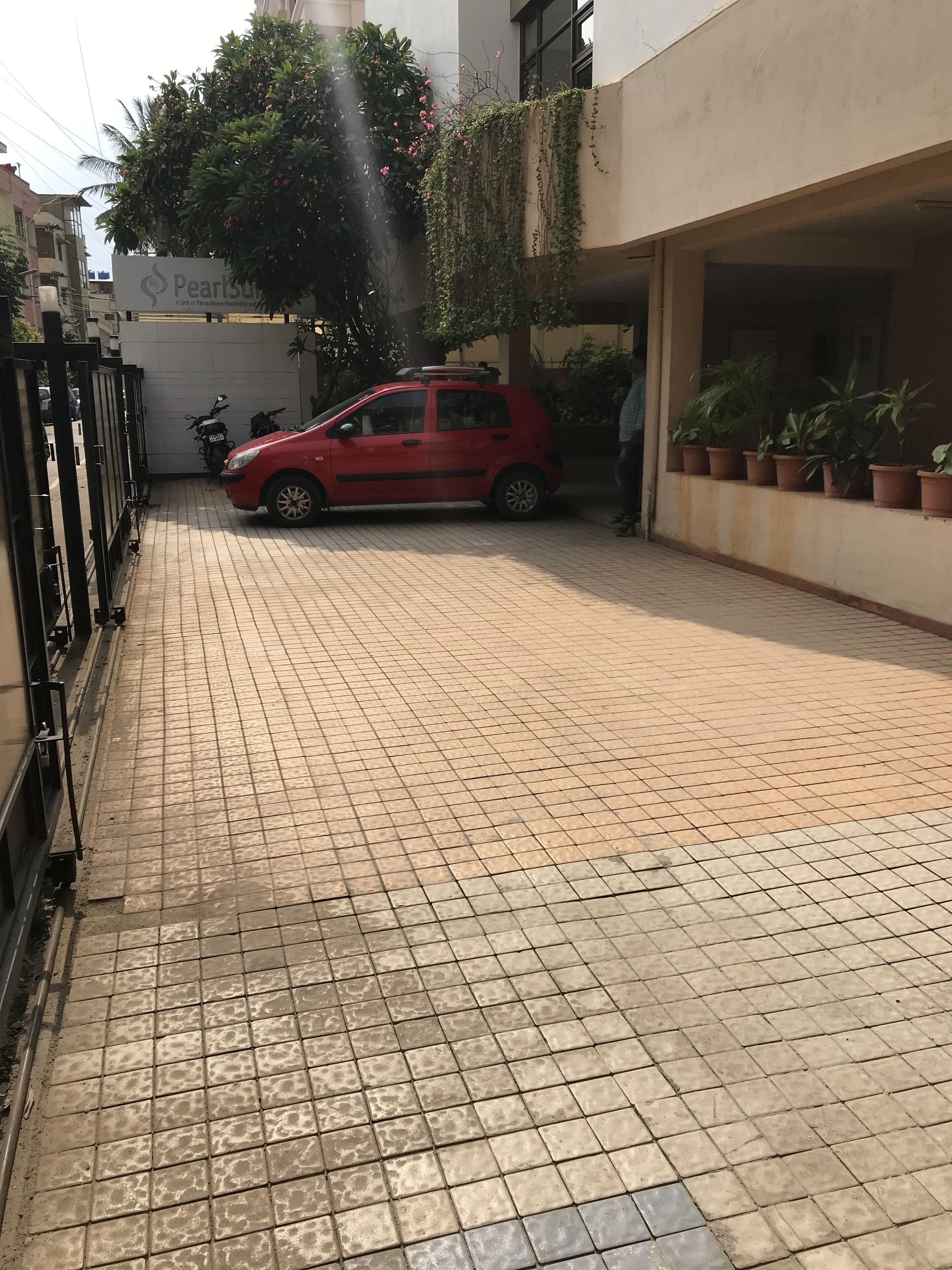 A paved courtyard with a red car parked near a gated entrance, surrounded by potted plants and a building with a sign.