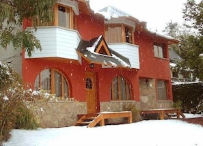 A red two-story house with a stone facade, wooden balcony, and a stone entrance. - Bungalows Maniu (San Carlos de Bariloche)