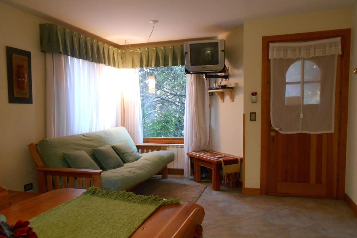 Una sala de estar acogedora con un sofá, un televisor fijado a la pared, una mesita y una puerta con ventanal.