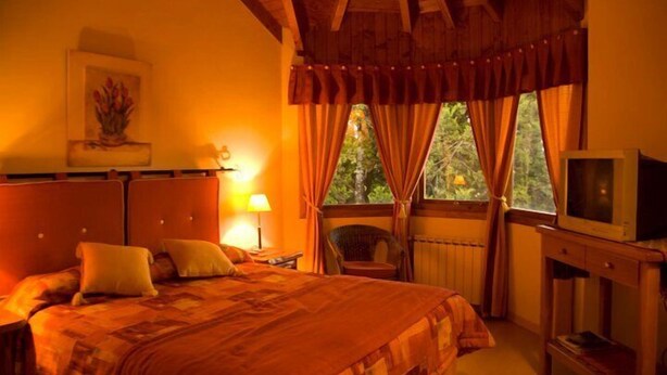 Un dormitorio con una cama grande, un televisor sobre un soporte de madera, una silla y una ventana con cortinas.