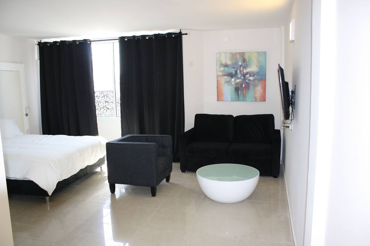 A modern hotel room with a bed, a black armchair, a black sofa, a white coffee table, and a painting on the wall.