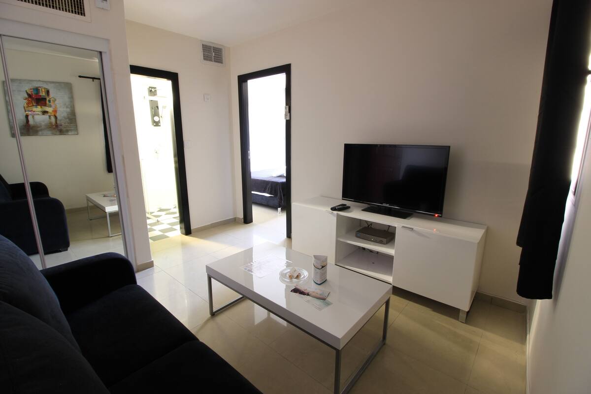 A modern living room with a flat-screen TV, a white coffee table, and a sofa. There are two doors leading to other rooms.