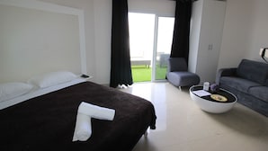Moderni hotellihuone, jossa on suuri sänky, harmaa sohva, pieni pyöreä sohvapöytä ja liukuovi, joka johtaa parvekkeelle.