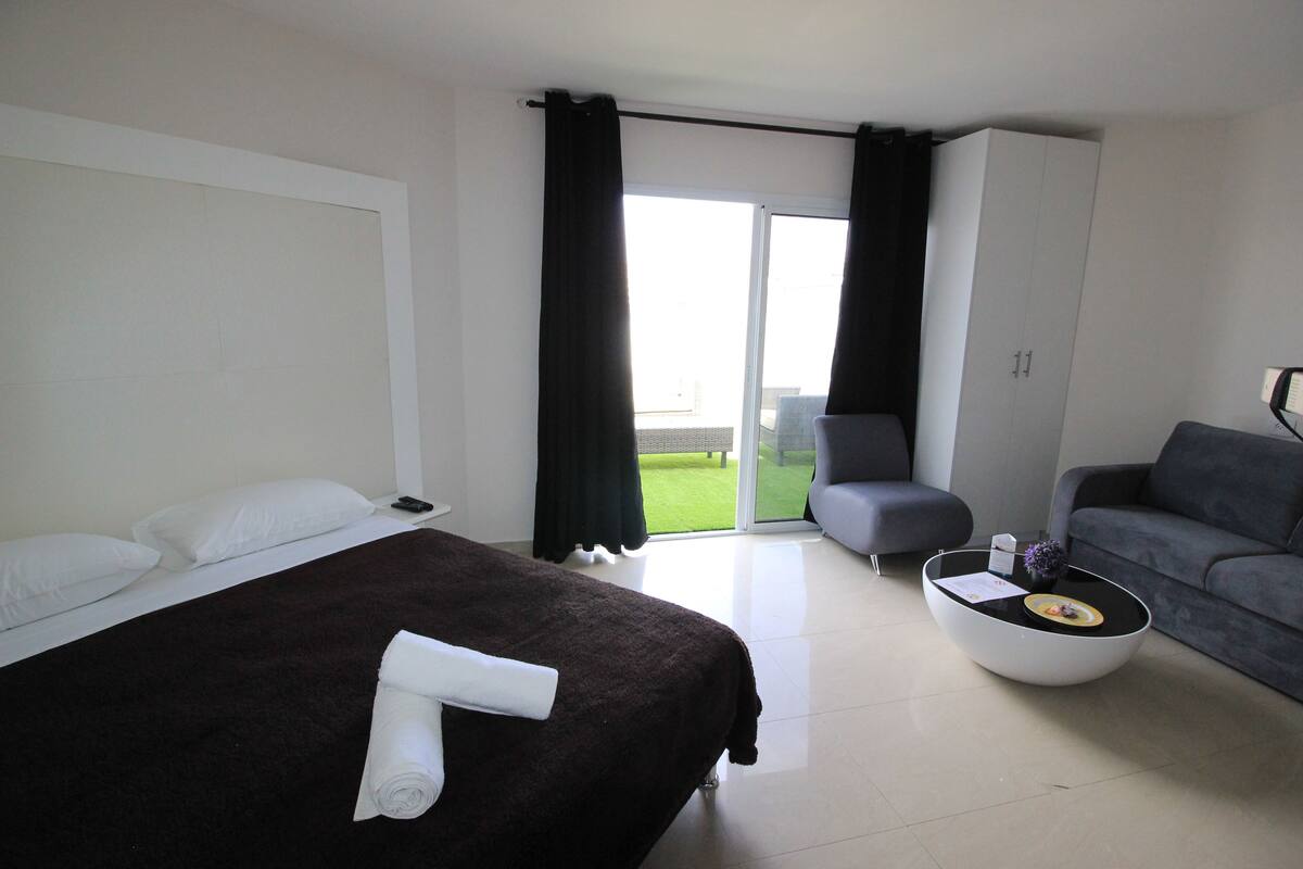 A modern hotel room with a large bed, a grey sofa, a small round coffee table, and a sliding glass door leading to a balcony.