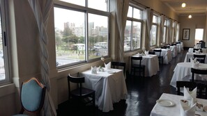 A restaurant with multiple tables set for dining, large windows with curtains, and a view of the cityscape.